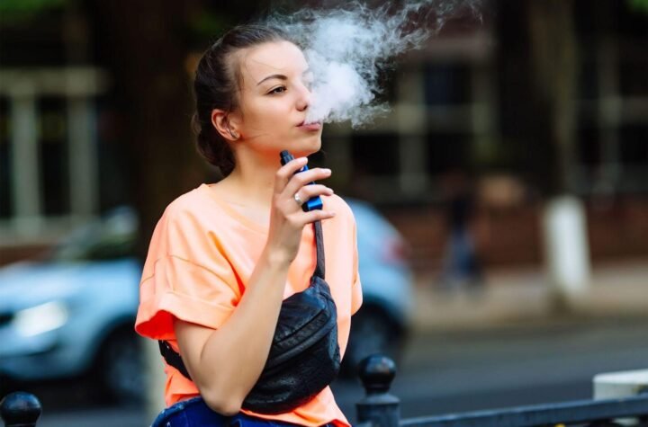 Girl in an orange shirt exhaling vape instead of bong from an electronic cigarette outdoors.